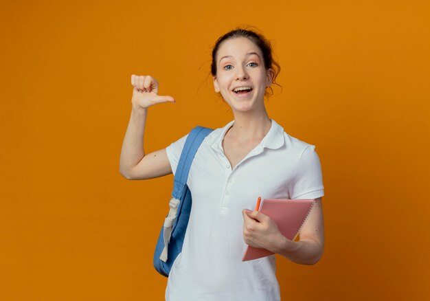Pod wrażeniem młoda ładna studentka noszenie plecaka trzymając notes i pióro, wskazując na siebie na białym tle na pomarańczowym tle z miejsca na kopię