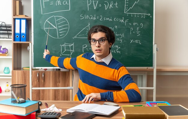 pod wrażeniem młoda nauczycielka geometrii kaukaskiej w okularach siedząca przy biurku z przyborami szkolnymi w klasie trzymająca rękę na biurku patrząca na przód wskazująca kijem na tablicę