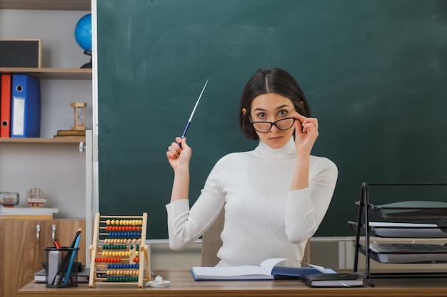 Bezpłatne zdjęcie pod wrażeniem młoda nauczycielka nosząca okulary wskazuje ze wskaźnikiem na tablicy siedzącej przy biurku z narzędziami szkolnymi włączonymi w klasie