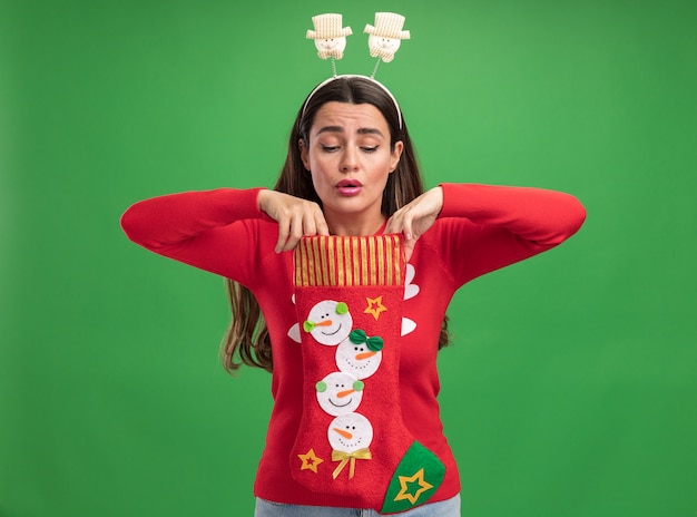 Pod wrażeniem młoda piękna dziewczyna ubrana w świąteczny sweter z świątecznym obręczem do włosów, trzymając i patrząc w świąteczne skarpetki na białym tle na zielonym tle