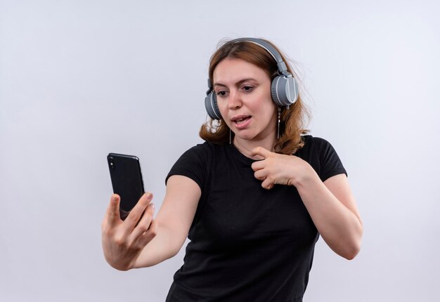 Pod wrażeniem młoda przypadkowa kobieta nosi słuchawki i trzyma telefon komórkowy na odosobnionej białej przestrzeni