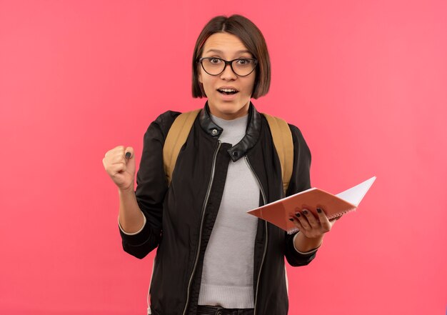 Pod wrażeniem młoda studentka w okularach i torbie z powrotem trzymając notes zaciskając pięść na różowym tle