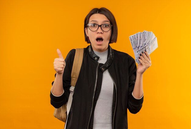 Pod wrażeniem młoda studentka w okularach iz powrotem worek trzymając pieniądze pokazując kciuk do góry na białym tle na pomarańczowo