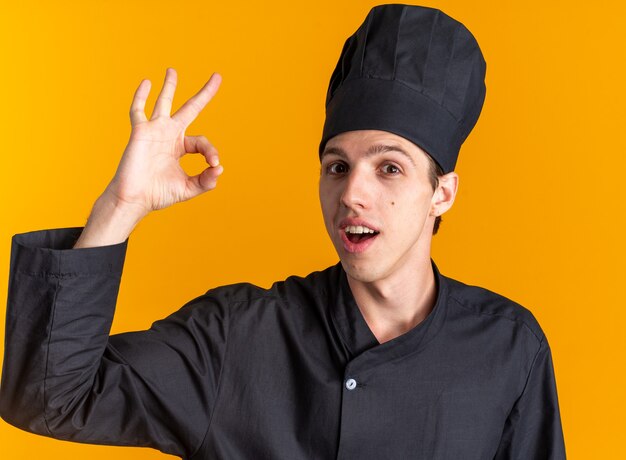 Pod wrażeniem młodego blond kucharza w mundurze szefa kuchni i czapce, patrząc na kamerę robi ok znak na pomarańczowej ścianie