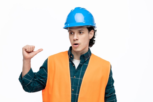 Bezpłatne zdjęcie pod wrażeniem młodego inżyniera płci męskiej w kasku i kamizelce ochronnej, patrząc na bok wskazujący za na białym tle