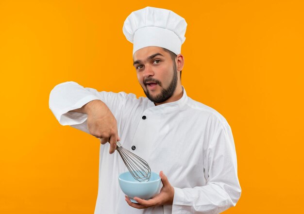 Pod wrażeniem młodego kucharza w mundurze szefa kuchni, trzymającego trzepaczkę i miskę na pomarańczowej ścianie