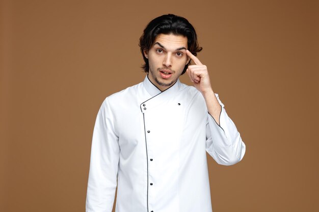 pod wrażeniem młodego szefa kuchni płci męskiej w mundurze, patrząc na kamerę pokazującą gest myślenia na białym tle na brązowym tle
