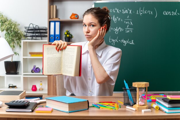 Pod wrażeniem młodej blondynki nauczycielka matematyki siedząca przy biurku z szkolnymi narzędziami, patrząca na kamerę pokazującą książkę do kamery trzymającą rękę na twarzy w klasie