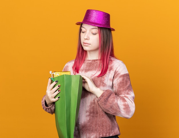 Bezpłatne zdjęcie pod wrażeniem młodej pięknej kobiety noszącej imprezowy kapelusz trzymający i patrzący na torbę z prezentami odizolowaną na pomarańczowej ścianie