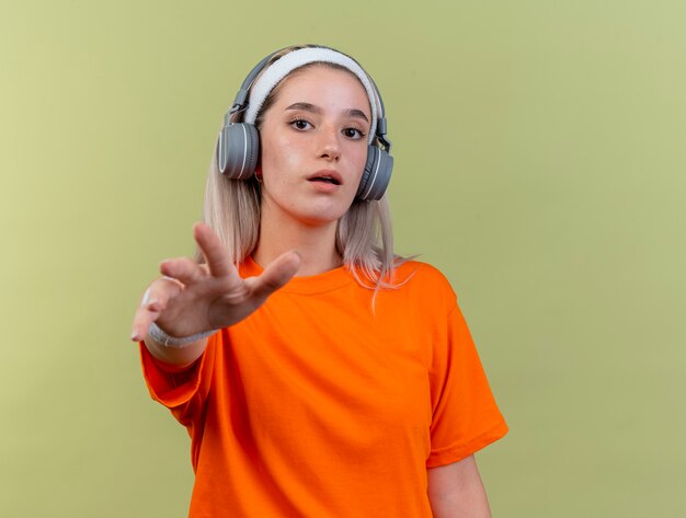 Pod wrażeniem młodej sportowej dziewczyny rasy kaukaskiej z szelkami na słuchawkach, noszącej opaskę na głowę i opaski na nadgarstkach wyciągające rękę