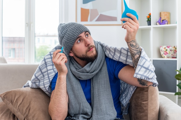 Bezpłatne zdjęcie pod wrażeniem młody chory mężczyzna ubrany w szalik i zimową czapkę siedzący na kanapie w salonie owinięty w koc trzymający lewatywę podnoszący jednego patrzącego na niego