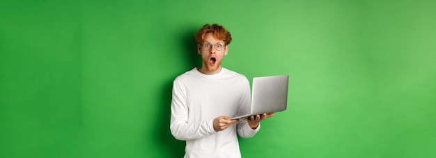 Bezpłatne zdjęcie pod wrażeniem młody człowiek w okularach opada szczęka i gapi się zdumiony po przeczytaniu promocji na laptopie stojącym ov