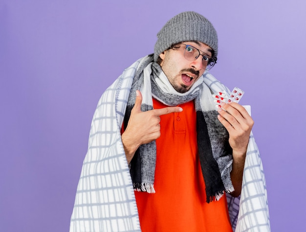 Bezpłatne zdjęcie pod wrażeniem młody kaukaski chory mężczyzna w okularach czapka zimowa i szalik owinięty w kratę, trzymając i wskazując na paczki kapsułek odizolowane na fioletowej ścianie z miejscem na kopię