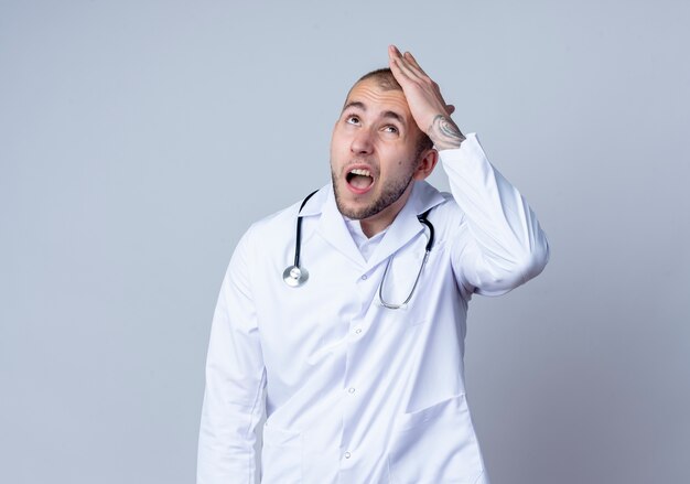 Pod wrażeniem młody lekarz płci męskiej ubrany w szlafrok i stetoskop wokół szyi, kładąc rękę na głowie i patrząc w górę na białym tle