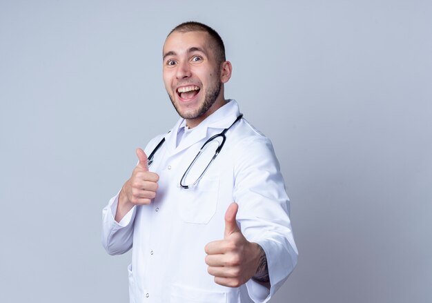 Pod wrażeniem młody lekarz płci męskiej ubrany w szlafrok i stetoskop wokół szyi pokazując kciuk do góry na białym tle