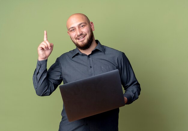 Pod wrażeniem młody łysy mężczyzna call center trzymając laptop i podnosząc palec na białym tle oliwkowej zieleni z miejsca na kopię
