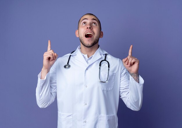 Pod wrażeniem młody mężczyzna lekarz ubrany w szlafrok i stetoskop patrząc i wskazując w górę na białym tle na fioletowo