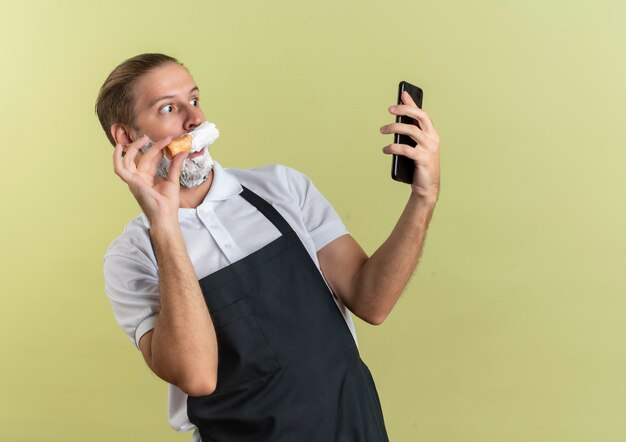 Pod Wrażeniem Młody Przystojny Fryzjer W Mundurze, Trzymając I Patrząc Na Telefon Komórkowy Oraz Nakładając Krem Do Golenia Pędzlem Do Golenia Na Własną Brodę Odizolowaną Na Oliwkowej Zieleni Z Miejscem Na Kopię