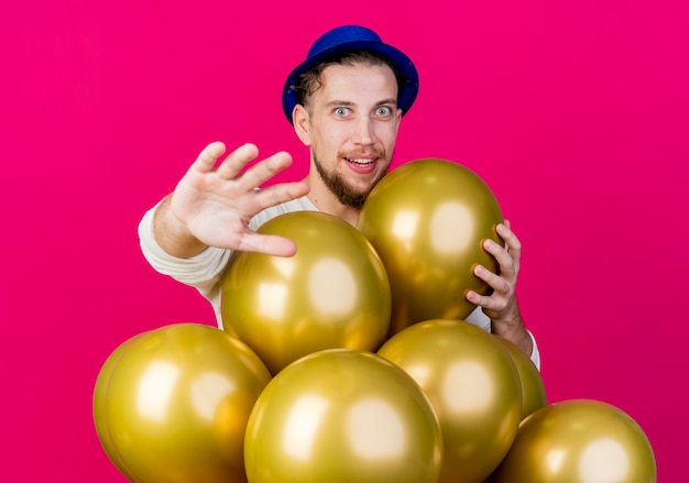 Pod wrażeniem młody przystojny słowiański facet w kapeluszu imprezowym stojący za balonami, kładąc rękę na balonie wyciągając rękę i patrząc na kamerę odizolowaną na szkarłatnym tle