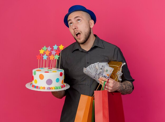 Pod wrażeniem młody przystojny słowiański imprezowicz w kapeluszu imprezowym, trzymając tort urodzinowy pieniądze pakiet prezentów i papierowe torby patrząc w górę na białym tle na szkarłatnym tle