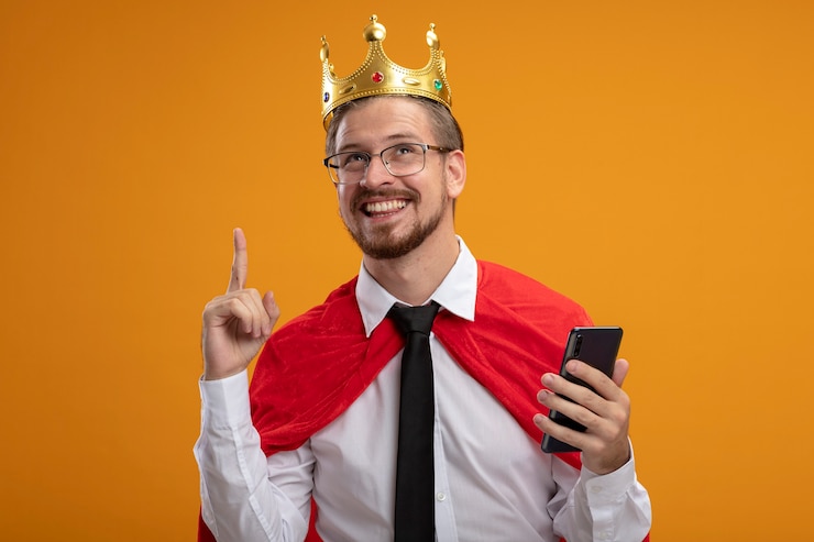 Bezpłatny plik Zdjęcie pod wrażeniem młody superbohater facet ubrany w krawat i koronę w okularach trzyma telefon i wskazuje na górę na białym tle na pomarańczowym tle