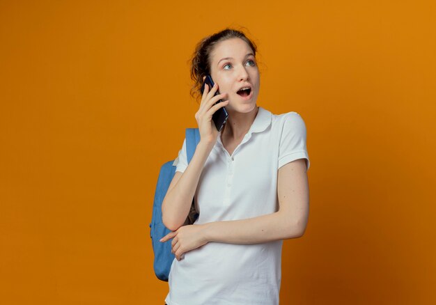Pod wrażeniem młodych ładnych studentek na sobie tylną torbę patrząc na bok rozmawia przez telefon na białym tle na pomarańczowym tle