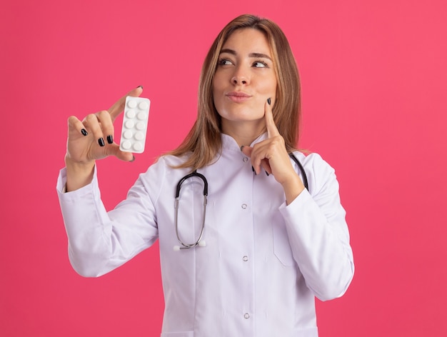 Pod wrażeniem patrząc z boku młoda kobieta lekarz ubrana w szlafrok medyczny ze stetoskopem trzymająca pigułki kładąc palec na policzku odizolowana na różowej ścianie