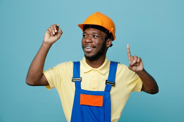 Pod wrażeniem punktów na młodym afroamerykańskim budowniczym w mundurze trzymającym znacznik na białym tle na niebieskim tle