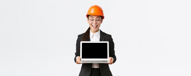Pod wrażeniem szczęśliwa azjatycka główna inżynierka pokazująca wspaniałe wyniki przedsiębiorstwa inwestorom stojącym w kasku ochronnym i garniturze, trzymająca laptopa i patrzącego z zadowoleniem na ekran komputera