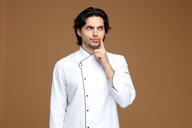 podejrzany młody mężczyzna szef kuchni ubrany w mundur, dotykając twarzy, patrząc na bok na białym tle na brązowym tle