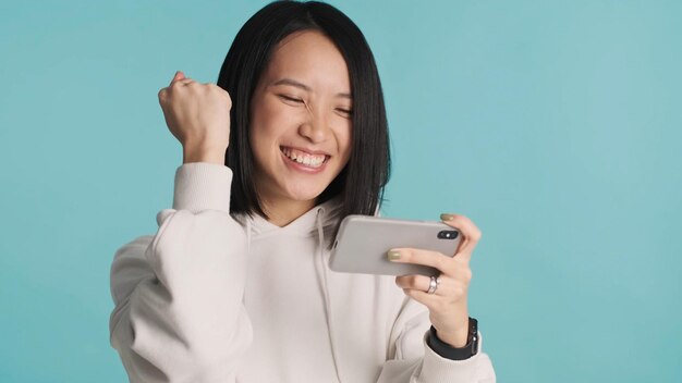 Podekscytowana Azjatka emocjonalnie grająca na telefonie komórkowym świętująca wygraną w grze online na białym tle na niebieskim tle