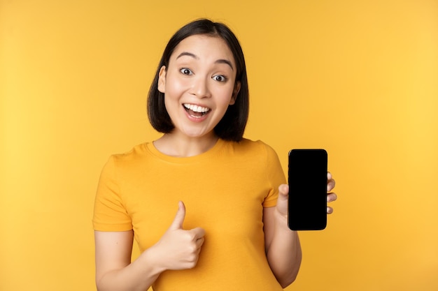 Podekscytowana Azjatka pokazuje kciuki na ekranie telefonu komórkowego, jak coś dobrego, polecając aplikację na smartfona stojącą na żółtym tle