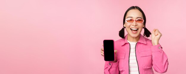 Podekscytowana Azjatka śmieje się i uśmiecha, pokazuje aplikację smartfona na ekranie telefonu komórkowego stojącą na różowym tle