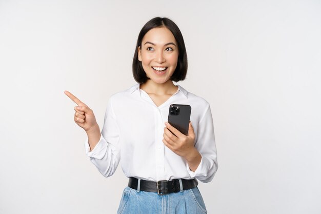 Podekscytowana azjatycka dziewczyna robi zakupy w aplikacji mobilnej, trzymając telefon i wskazując palcem w lewo na pustej przestrzeni, pokazując tekst promocyjny stojący ze smartfonem na białym tle