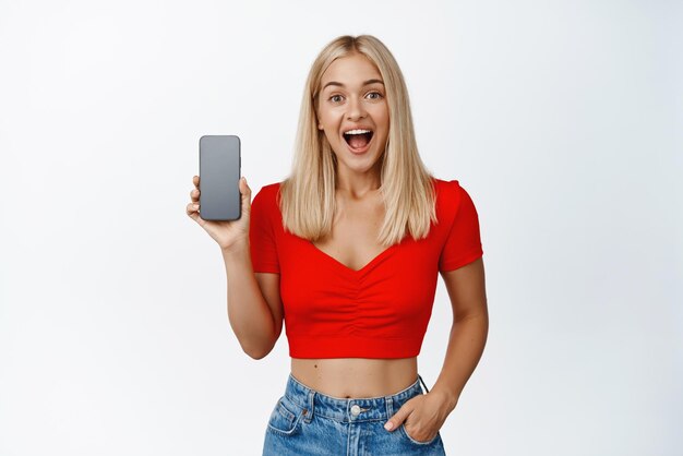 Podekscytowana blond dziewczyna pokazująca interfejs aplikacji na ekranie telefonu komórkowego lub aplikację zakupową, uśmiechając się zdumiona, stojąc na białym tle