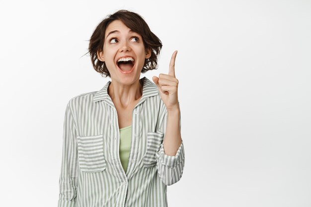Podekscytowana brunetka dziewczyna śmiejąca się, uśmiechnięta i wskazująca palcem w górę, chichocząca nad śmieszną reklamą, stojąca w koszuli na białym tle.