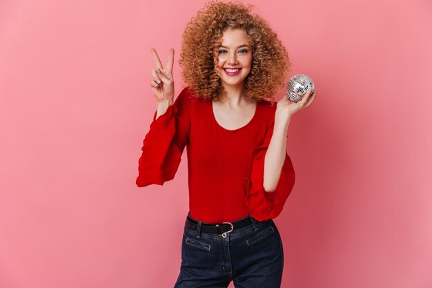Podekscytowana dziewczyna ubrana w czerwoną bluzkę i dżinsy pozuje z kulą dyskotekową i pokazuje znak pokoju na różowym tle