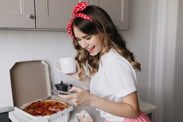 Podekscytowana Dziewczyna Ze Wstążką We Włosach Picia Kawy Rano. Kryty Ujęcie Uroczej Pani Jedzącej Pizzę Podczas śniadania.