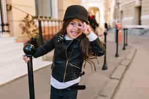 Bezpłatne zdjęcie podekscytowana dziewczynka ubrana w skórzaną kurtkę i pasek trzymający żelazny filar i pozująca ze znakiem pokoju na tle miasta.