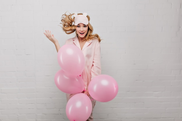 Podekscytowana jasnowłosa dziewczyna w różowej piżamie trzymająca balony z helem. Wewnątrz portret inspirowanej młodej damy w modnej masce do spania tańczącej na jasnej ścianie.