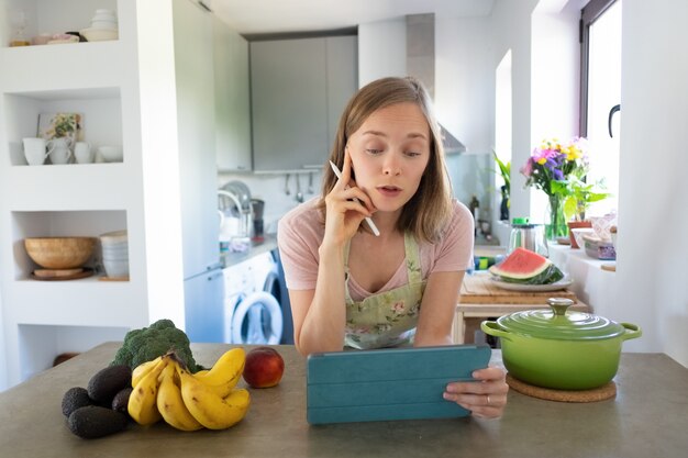 Podekscytowana kobieta ogląda lekcje gotowania online w swojej kuchni, opierając się o stół, używając tabletu w pobliżu rondla i świeżych owoców na blacie. Przedni widok. Gotowanie w domu i koncepcja zdrowego odżywiania