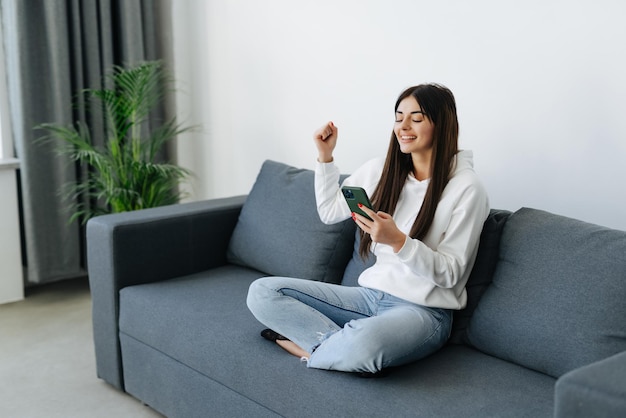 Podekscytowana kobieta oglądająca treści multimedialne on-line w telefonie komórkowym, siedząca na kanapie w salonie w domu