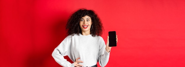 Podekscytowana Kobieta Z Kręconymi Włosami I Czerwonymi Ustami, Pokazująca Pusty Ekran Smartfona I Krzycząca Z Radości St
