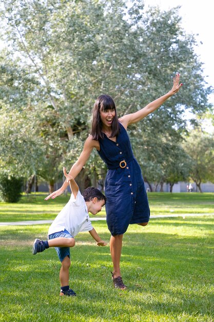 Podekscytowana mama i synek grają w aktywne gry na świeżym powietrzu, stojąc i balansując na jednej nodze, wykonując zabawne ćwiczenia w parku. Rodzinna koncepcja aktywności i wypoczynku na świeżym powietrzu