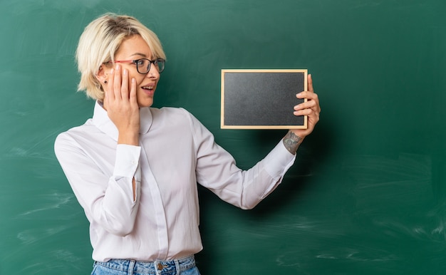 Podekscytowana młoda blondynka nauczycielka w okularach w klasie stojąca przed tablicą pokazującą mini tablicę patrzącą na nią trzymając rękę na twarzy z kopią miejsca