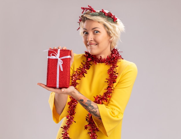 Podekscytowana młoda blondynka ubrana w świąteczny wieniec na głowę i świecącą girlandę na szyi trzyma pakiet prezentów bożonarodzeniowych, patrząc gryzącą wargę na białej ścianie z miejscem na kopię