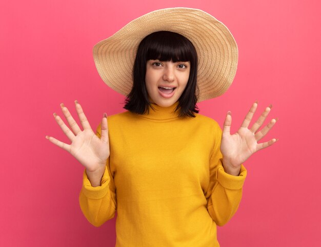 Podekscytowana młoda brunetka kaukaska dziewczyna w kapeluszu plażowym z uniesionymi rękami