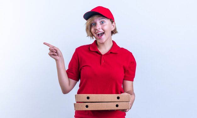 podekscytowana młoda dostawa dziewczyna ubrana w mundur i czapkę, trzymająca pudełka po pizzy i punkty z boku na białym tle na białej ścianie z miejscem na kopię