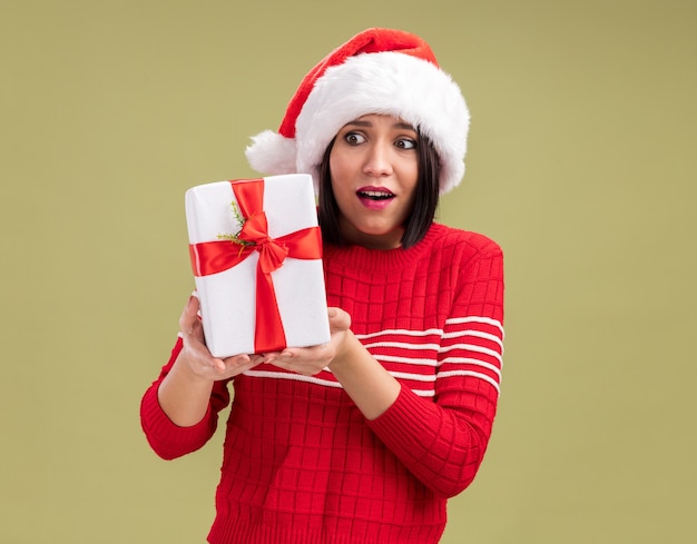 Podekscytowana młoda dziewczyna ubrana w santa hat trzyma i patrząc na pakiet prezentów na białym tle na oliwkowym tle z miejsca na kopię