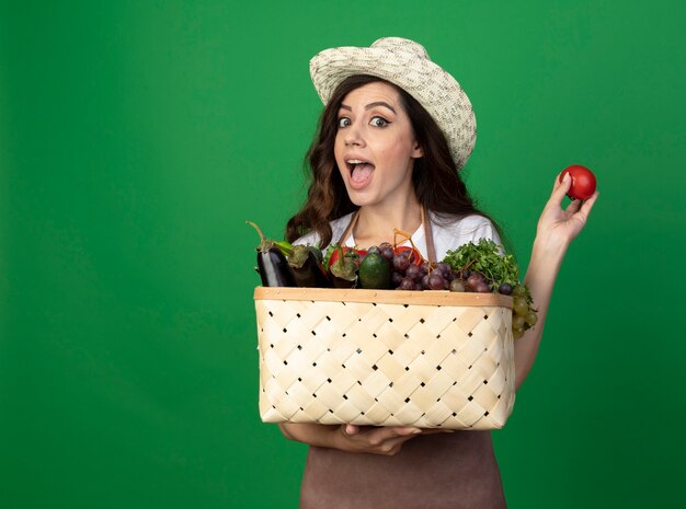 Podekscytowana młoda kobieta ogrodniczka w mundurze na sobie kapelusz ogrodniczy trzyma kosz warzyw i pomidora na białym tle na zielonej ścianie
